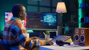 Afro-amerikan bilişim uzmanı makine öğrenimi için yapay zeka sistemini yönetiyor, terminal penceresinde kodlama yazılımı var. Gelecekteki arayüz programı siber güvenliğe odaklanır, veritabanı için ağ oluşturur. Kamera A.