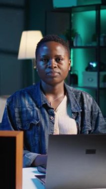 Gecenin geç saatlerinde neon ışıklı apartmandaki ofis masasında oturan neşeli kadının dikey video portresi. RGB 'deki neşeli insan oturma odasını aydınlatıyor. Boş zamanların tadını çıkarıyor. Kamera B.