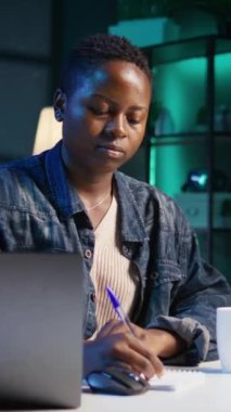 Dikey video, Jolly Freelancer telgraf çekerken kalemle fikirleri not alıyor. Ev ofis masasında çalışan neşeli bir çalışan, dizüstü bilgisayardan bilgi okuyor ve deftere yazıyor.