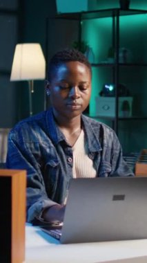 Ev ofisinin masasında oturmuş laptoptaki görevleri çözmeye hazırlanan neşeli bir kadının dikey video portresi. Dijital cihazın önünde neşeli bir serbest yazar, oturma odasında e-posta iletişimi yapıyor, kamera.