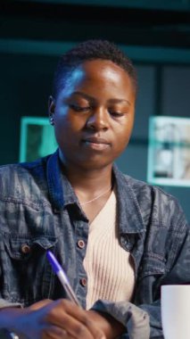 Ofis masasındaki sandalyeye oturup not defterine notlar yazan gülümseyen bir kadının dikey video portresi. Oturma odasında keyifli bir insan. Bilgisayardan gelen bilgileri kağıda döküyor. Kamera B.