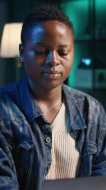 Masada dizüstü bilgisayarda yazı yazan, görevleri çözen gülümseyen Afrikalı Amerikalı kadının dikey video portresi. Oturma odasından iş ortaklarına e-posta göndermek için dizüstü bilgisayar kullanan neşeli bir çalışan.