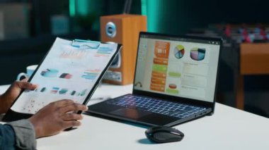 Ofis masasında oturan bir kadın, iş belgelerine bakıyor. Panodaki finansal çizelgeleri okurken, oturma odasında not defteri kullanan Afrikalı Amerikalı serbest yazar, kamera A