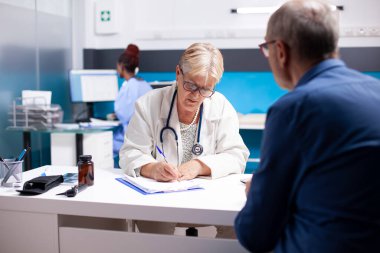 Kıdemli doktor, hastanede sağlık kontrolü sırasında hasta erkek emekliye ait hastalık belirtileri yazıyor. Yaşlı bir kadın doktor klinikte yaşlı bir adamla görüşürken pano kullanıyor..