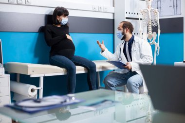 Doktor panoyu tutuyor ve müstakbel anne adayıyla klinik ziyareti sırasında tıbbi tedavisi hakkında sabırla konuşuyor. Hastanede hamile bir kadınla doğum öncesi bakım detaylarını açıklayan bir doktor..