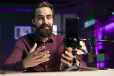 ASMR içerik yaratıcısı, stres karşıtı videolar yapmak için tripodun üzerinde kaliteli mikrofon ve cep telefonu kullanıyor, dinleyicileri yatıştırmak için fısıldayan sesi kullanıyor. Seyircileri uyutmak için profesyonel mikrofon kullanan bir adam