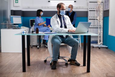 Male doctor wearing lab coat and face mask, sitting at desk with device, focused on preparing for clinical consultations. Physician working on laptop as nurse consults with old patient in background. clipart