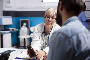 Klinikte sakallı bir adam emekli bir kadın doktor olarak oturur ve reçeteli ilacın etkilerini açıklar. Yaşlı doktora yakın çekim, hasta hastaya bir şişe ilaç sunuyor..