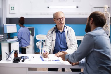 Laboratuvar önlüklü yaşlı doktor sakallı erkek hastanın endişelerini dikkatle dinliyor ve hastane ofisindeki panoya yazıyor. Yaşlı hekim klinikte hasta genç bir adamla tıbbi detayları inceliyor..