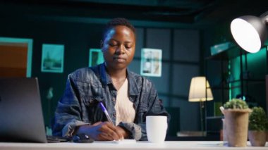 Ofis masasında oturan gülümseyen bir kadının portresi. Not defterine notlar yazıyor. Oturma odasında keyifli bir insan. Bilgisayardan gelen bilgileri kağıda döküyor. Kamera B.