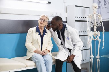 African american doctor examining the ear of elderly white woman while seated on hospital bed. Professional physician holding otoscope performs medical examination on senior female patient. clipart