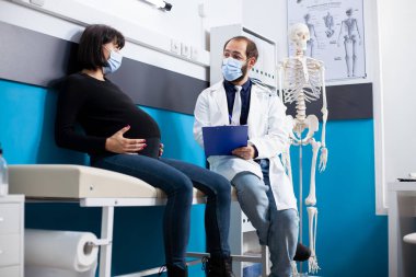 Genç sağlık uzmanları pano tutuyor ve klinikte teşhis detaylarını tartışarak hamile hastalara danışıyor. Erkek doktor hasta yatağında hamile annesinin yanında oturup annelik tavsiyesi veriyor..