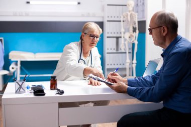 Dostane kıdemli doktor panodaki tıbbi sonuçlara işaret ederek emekli erkek hastaya rehberlik ediyor. Emekli adam sağlık bilgilerini yaşlı kadın hekimin yardımıyla yazıyor..