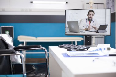 Uzaktan tanı koymak için hazırlanmış boş bir hastane odası ve bilgisayar ekranında görüntülü bir doktor var. Sağlık hizmetleri iletişimine modern yaklaşımı yansıtan işlenmemiş klinik ofisi.
