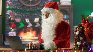 Saint Nicholas is busy online browsing Christmas websites in his festive North Pole cabin. Santa making preparations on internet webpages, spreading the magical winter holiday season. Camera A. clipart