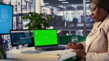 Genç genel müdür laptopta yeşil ekran kullanıyor ve personeli için performans ölçümlerini değerlendiriyor, bir planlama oturumu düzenliyor. Afrikalı Amerikalı CEO liderlik becerileri gösteriyor. Kamera B.