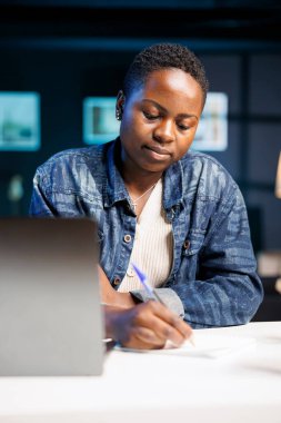 Genç siyahi bir kadın, teknoloji kullanarak müşterileri araştırıyor ve onlarla bağlantı kuruyor. Not defterine yazmak için kalem kullanan Afrikalı Amerikalı serbest yazar, çevrimiçi çalışmalarında üretken ve yenilikçi.