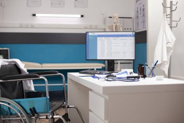 Tekerlekli sandalyedeki boş masadaki bilgisayar monitörü yaklaşan hasta muayenelerini gösteriyor. Sağlık hizmetleri için hastane teçhizatı ile parlak, temiz klinik alanı.