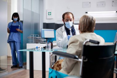 Sıhhiye uzmanı, klinikteki tekerlekli sandalyedeki yaşlı kadına tedavi seçeneklerini açıkladı. Hasta emekliye tıbbi danışmanlık yapan erkek doktor, arka planda pano taşıyan hemşire olarak....