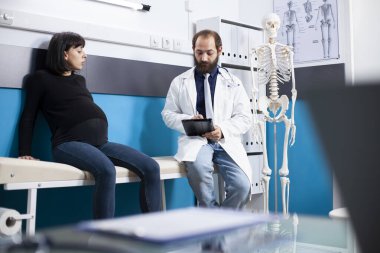 Hamilelik kontrolü sırasında, dijital cihazlı erkek doktor klinikte yanında oturan müstakbel kadına tedavi seçeneklerini açıklıyor. Doktor, hamile bir hastayla tabletteki tıbbi sonuçları gözden geçiriyor..