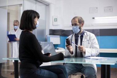 Erkek doktor, kadın hastaya tıbbi konsültasyon sırasında yüz maskesi takan ilaçları açıklıyor. Doktor ilaç şişesini tutuyor ve genç beyaz bir kadının tedavisi için ilaç etkisini tartışıyor..