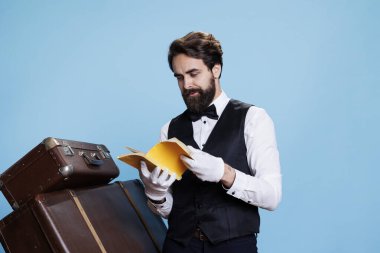 Doorman reads literature fragments from book, enjoying hobby against blue background in studio. Hotel concierge worker recites poetry or novel tales, man wearing formal suit and tie. clipart