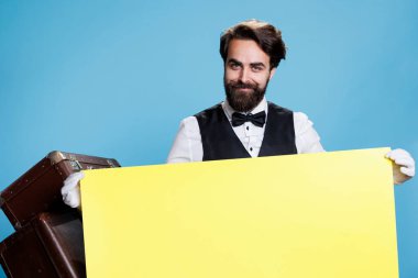 Young bellboy holds yellow blank poster against blue background, professional hotel employee creating marketing advertisement with empty banner sign. Bellhop porter with cardboard. clipart