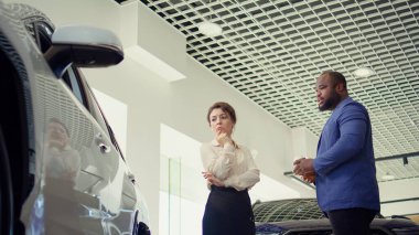 Araba galerisinde yürüyen müşteriler mevcut modellere göz atıyorlar ve ideal yatırım seçeneğini seçmeye çalışıyorlar. Galeride araç almak isteyen karı koca, kamera B