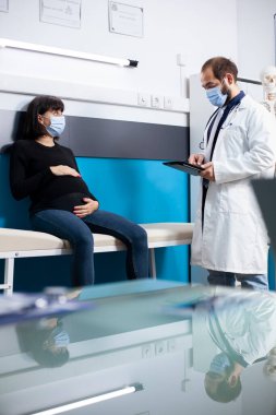 Kadın hastaya hamilelik tavsiyesi vermek için tablet kullanan erkek doktor. Genç hamile kadın hastane yatağında oturuyor ve doktordan doğum öncesi tedavi görüyor.