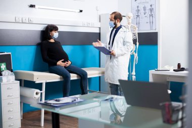 Tıp ofisinde, laboratuvar önlüklü bir doktor hamile bir kadının yanında duruyor ve ona sağlık raporu veriyor. Erkek jinekolog, genç hamile annesine danışmanlık yapıyor..