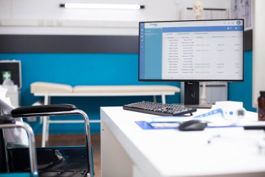 Boş tekerlekli sandalye kullanıcı masası ve gelecek randevular için hasta sağlık kayıtlarını gösteren bilgisayar ekranı. Muayene araçları ve organize tıbbi belgelerle klinik ayarları.