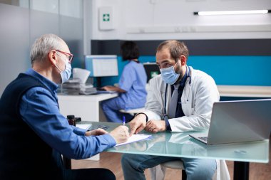 Pratisyen hekim, yaşlı beyaz bir adama panoda tıbbi bilgiler doldurmasında yardımcı oluyor. Genç doktor ve erkek emekli, her ikisi de maske takıyor, klinikte sağlık değerlendirmesini tartışıyorlar..