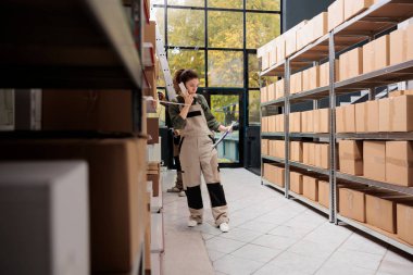 Depo çalışanı mesai saatinde sabit telefon şirketinin sahibi ile envanter detaylarını tartışıyor. Müşteri emirleriyle çalışan bir denetçi, depoda nakliye için ürünler hazırlıyor.