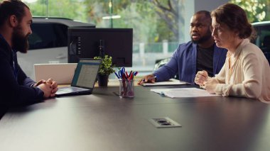 Satış temsilcisi, müşteriler için araba test sürüşü planlamak için dizüstü bilgisayardaki randevu listesini inceliyor. Showroom temsilcisi müşterilerle tartışıyor, test sürüşünü düzenliyor, kamera B