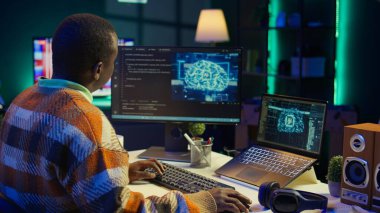 Afro-amerikan bilişim uzmanı makine öğrenimi için yapay zeka sistemini yönetiyor, terminal penceresinde kodlama yazılımı var. Gelecekteki arayüz programı siber güvenliğe odaklanır, veritabanı için ağ oluşturur. Kamera B.