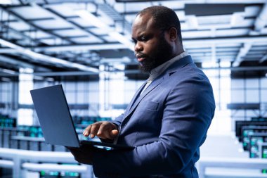 Sunucu odasındaki mühendis yeni sipariş edilen depolama birimlerini dizüstü bilgisayarla kuruyor. Veri merkezi depolama tesisinde çalışan Afrikalı Amerikalı bir adam.