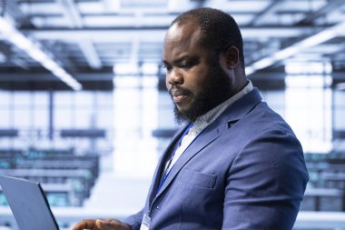 Veri merkezindeki bilgisayar bilimcisi laptop kullanarak veri kaybından korunmak için yedek çözümler geliştiriyor ve verimliliği sağlıyor. Sunucu odasında teknik sorunları çözen Afrikalı Amerikalı bir adam var.