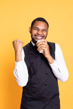 Happy confident waiter drinking a cup of coffee and feeling pleased, working in customer focused catering industry. Cheerful restaurant employee enjoying caffeine drink on duty. clipart