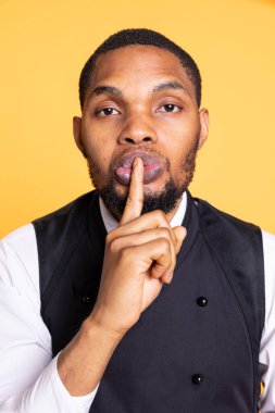 Five star restaurant staff showing the silence symbol and asking for secrecy, presenting the mute sign against yellow background. Waiter in apron asking to keep secret and be quiet. clipart