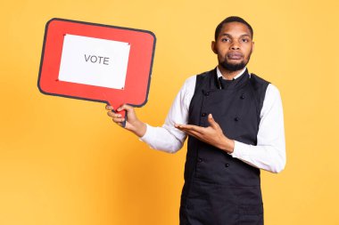 Waiter does an advertisement for politics and showing a speech bubble with the message to vote, standing against yellow background. Restaurant staff holds a cardboard sign. clipart