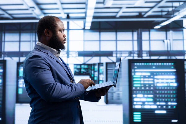 Programmer using laptop in data center with advanced technology, ensuring cybersecurity and efficient network management. Professional in server hub optimizing data center infrastructure with notebook