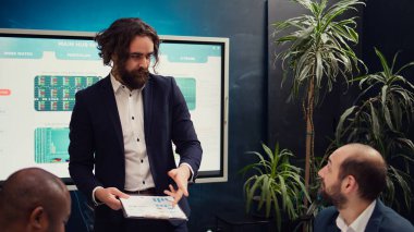 Takım lideri, şirket brifinginde çalışma verimliliği ve optimizasyonu tartışmak için son performans ölçümleriyle panoları paylaşıyor. Müdür personeline danışıyor. Kamera B.