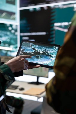 Erkek bilişim uzmanı, uydu dünya haritasına bağlı bir tablet kullanarak askeri karargah kullanarak askerlerin izini sürmeyi açıklıyor. Telekomünikasyon ekibi hava desteği sağlıyor. Kapat..