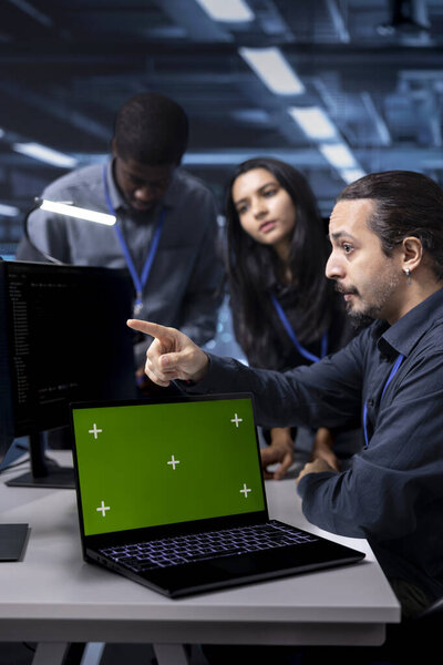 Multiethnic team of engineers in data center using system monitoring software on green screen laptop. Diverse coworkers in server hub repairing infrastructure with mockup notebook