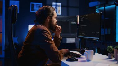 Neon lit ev ofisinde uzaktan iş değişimi sırasında web geliştirme programcısı, kodlama projesini bitiriyor. Bilgisayar masasında oturan bilişim uzmanı, masaüstü bilgisayarı, kamera B üzerinde çalışıyor.