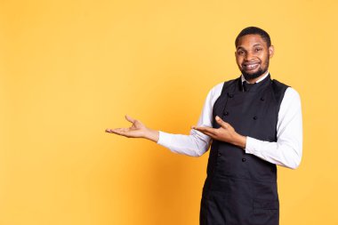 Afro-amerikan garson sağındaki ya da solundaki bir şeyi işaret ederek stüdyonun yan taraflarını gösteriyor. Müşteri odaklı çalışan arka planda üniforma ve önlük giyer.