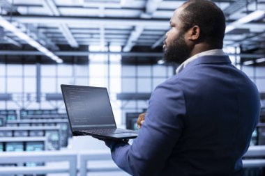 Veri merkezindeki sistem yöneticisi rackmontların en uygun şekilde çalıştığından emin oluyor. Sunucu merkezindeki Afrikalı Amerikalı adam süper bilgisayar platformlarında enerji tüketimini izlemek için dizüstü bilgisayar kullanıyor..