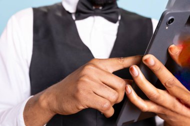 Professional bellhop browses hotel website on a tablet in studio, wearing suit and tie while he checks the room availability at five star resort. Doorman working in hospitality industry. clipart