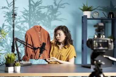 Medya yıldızı elinde turuncu ceketi tutuyor, geçenlerde aldığı hayranlarını gösteriyor, stüdyoda kendini filme çekiyor. Stüdyodaki içerik yaratıcısı seyirciye yeni alımlar sunar