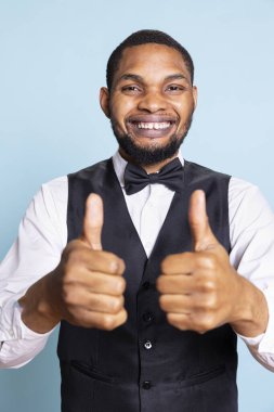 Smiling bellboy in suit and tie showing thumbs up gesture in studio, offering his approval and being satisfied. Positive doorkeeper gives a like sign, trust and professionalism. clipart